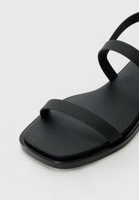 Black sandal with two thin straps and a square toe, featuring a smooth matte finish and a flat sole. Minimalist design and sleek lines.