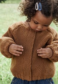 Brun fleecejakke med tekstureret, blød overflade og rundet halsudskæring. Synlige sømme og brede manchetter. Barnets hænder rører ved stoffet.