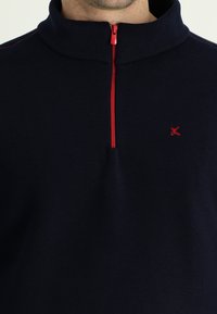 Marineblauwe fleece pullover met hoge kraag, voorzien van een rode rits en rode stikaccenten. Klein geborduurd logo op de borst.