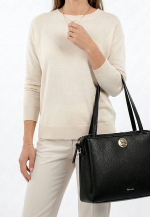 Vrouw in een crème trui en broek die een zwarte leren handtas met een gouden sluiting en dubbele handvatten aan haar rechterarm draagt.