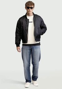 Jovem vestindo uma jaqueta bomber preta, óculos de sol, sweatshirt branca, jeans azuis e tênis brancos, posando contra um fundo simples.