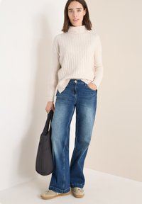 Lys beige ribbet sweater med høj hals, parret med blå vidde jeans. En simpel grå skuldertaske og beige sko fuldender outfittet.