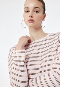 Lyserød langærmet bluse med vandrette brune striber, der har en bådudskæring og ribbede manchetter. Modellen har store hoops øreringe på.