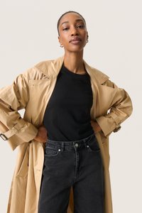 Beige trenchcoat over en sort t-shirt og mørke jeans. Frakken har en bred krave og justerbare manchetter. Glat stoftekstur.