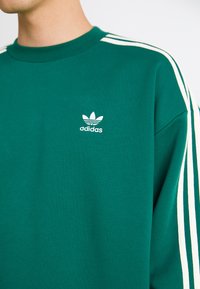 Sweater verde com o logo branco da Adidas, design de pescoço redondo e riscas brancas ao longo das mangas. Tecido macio e texturado.