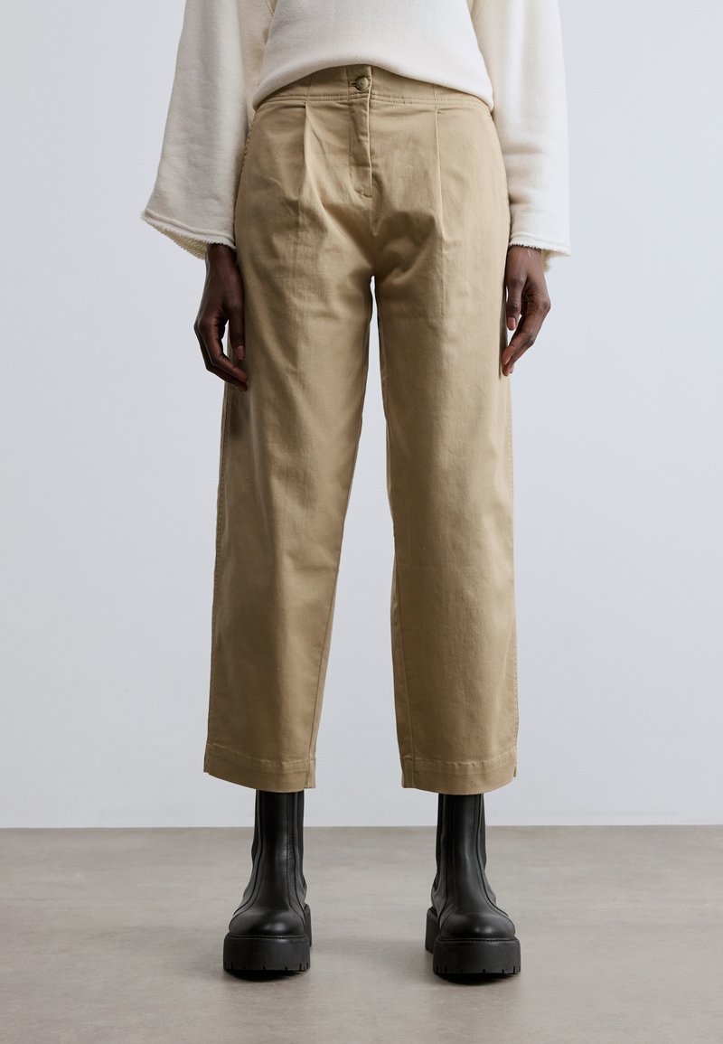 Bézs színű, pamutból készült, rövidített nadrág gombos záródással, elől gyűrött részletekkel és zsebekkel, fekete, vastag talpú csizmával párosítva.