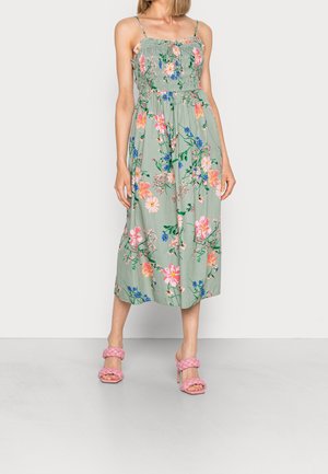 Floral maxi dress in light green. Features smocked bodice, thin straps, and vibrant flowers in pink and blue. Paired with pink heeled sandals.