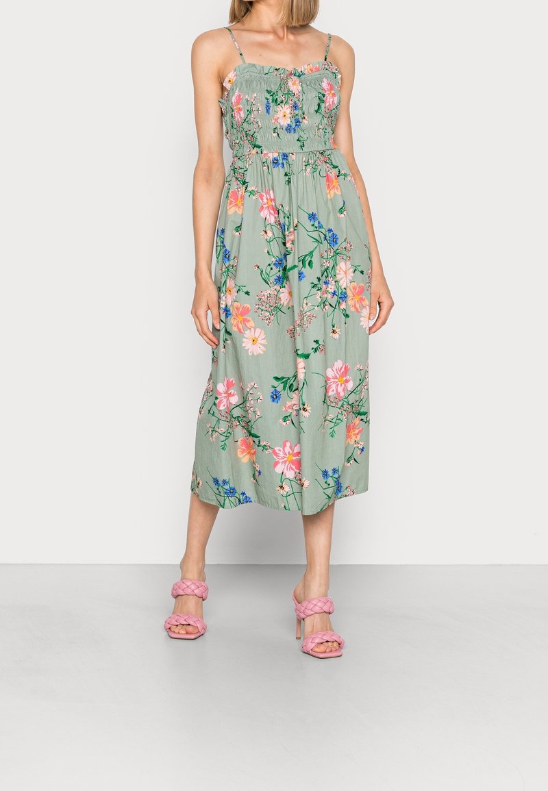 Robe maxi fleurie en vert clair. Caractérisée par un corsage froncé, des bretelles fines et des fleurs vibrantes en rose et bleu. Associée à des sandales à talons roses.