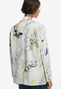 Veste blanche avec broderies florales colorées dans diverses teintes, dotée d'un revers et de boutons sur les poignets.