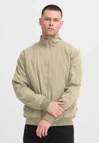 Veste bomber beige avec un col haut, fermeture éclair, poignets côtelés et une poche à zip sur la manche. Texture lisse avec un design minimaliste.