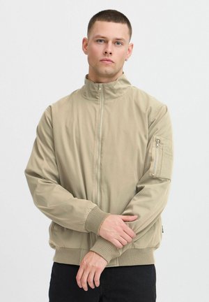 Khaki bomber jacket featuring a high collar, zip closure, ribbed cuffs, and a zippered sleeve pocket. Smooth texture with minimalistic design.