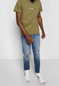 Olivově zelené bavlněné tričko s šedým logem "Calvin Klein Jeans", spárované s děravými světle modrými džíny a bílými teniskami se zlatými akcenty.