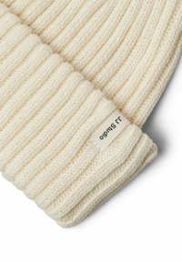 Gorro de punto color crema con canalé vertical, que presenta un dobladillo doblado y una pequeña etiqueta de tela blanca con texto en negro.