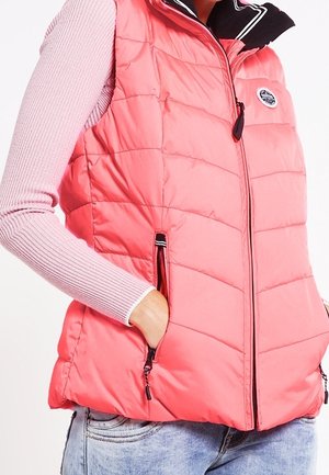 Person trägt eine korallenrosa gesteppte Weste über einem hellrosa gerippten Langarmshirt mit einer Hand in der Westentasche und hellblaue Jeans.