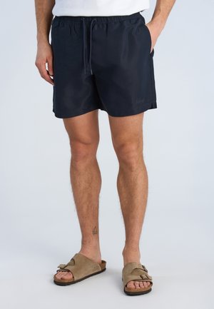 Mann trägt schwarze Shorts mit Kordelzug und beigen Schnallensandalen, steht mit den Händen in den Taschen vor einem hellgrauen Hintergrund.