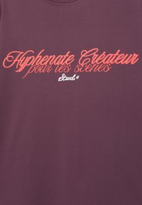 Mørkerød t-shirt med buet halsudskæring, der har hvid og orange trykt tekst, der læser "Hyphenate Createur pour les scènes" og logoet "Stwd."