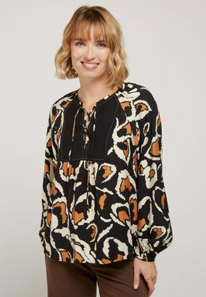 Blouse noire avec imprimé léopard orange et crème, comprenant un col en V avec des lacets, des manches longues et une coupe décontractée. Tissu texturé.