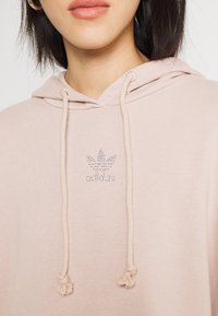 Helle rosa Kapuzenjacke aus weichem Stoff, mit verstellbarem Kapuzenzug und einem Strass-Adidas-Logo auf der Brust. Mit einer glatten Oberfläche.