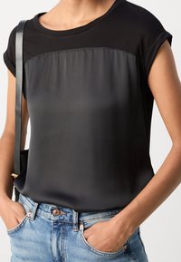 Kobieta nosząca czarną bluzkę z krótkim rękawem z satynowym przodem, niebieskie dżinsy oraz czarny pasek torby na ramieniu widoczny po lewej stronie.