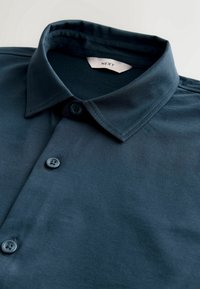 Chemise bleue foncée à boutons avec col et trois boutons visibles, posée à plat avec l'étiquette de la marque "NEXT" à l'intérieur du col.