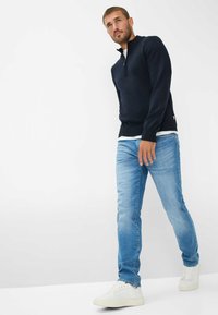 Maglione a collo zip navy sopra una camicia bianca, abbinato a jeans azzurri e sneakers bianche, indossato da un modello che cammina su uno sfondo neutro.