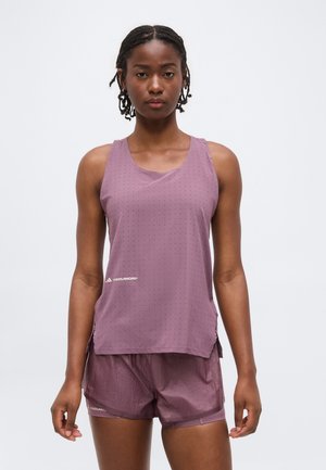 Γυναίκα που φοράει αμάνικο αθλητικό τοπ σε μωβ χρώμα και ταιριαστό σορτς, στέκεται μπροστά σε απλό φόντο.