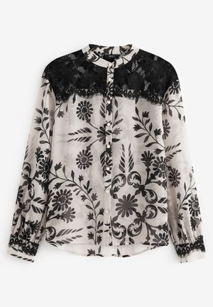 Sheer long-sleeve blouse with black floral patterns and black lace detail across upper back and cuffs, worn with black pants.