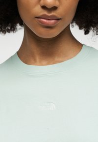 Κοντινό πλάνο ενός ατόμου που φοράει ανοιχτό πράσινο μπλουζάκι με στρογγυλή λαιμόκοψη και μικρό λευκό λογότυπο "The North Face" στο στήθος.