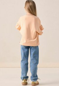 Kind dat met de rug naar de camera is gekeerd en een perzikkleurig topje met ruffles, blauwe jeans met kleine gele hartjespatronen, en beige sneakers met witte veters draagt.