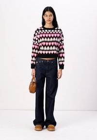 Svart strikket genser med rosa, hvite og grønne hjerter, høy midje på blå jeans, brune platåsko og en liten brun håndveske.