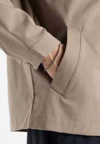 Beige jas met een ontspannen pasvorm, voorzien van een zijzak en een gladde textuur. Omgeslagen mouwen met subtiele stiksels. Zilveren armband zichtbaar.