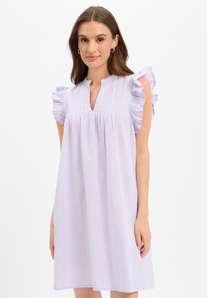 Frau trägt ein hellviolettes ärmelloses Kleid mit Rüschen an den Schultern und einem geteilten V-Ausschnitt, stehend vor einem weißen Hintergrund.