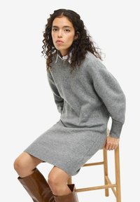 Vestito a maglia grigia con maniche lunghe, polsini a coste e tasche laterali, abbinato a stivali lucidi marroni fino al ginocchio su uno sgabello di legno.