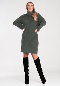 Knitdress in khakigrün mit hohem Kragen, langen Ärmeln und figurnaher Silhouette. Getragen mit knielangen schwarzen Wildlederstiefeln.