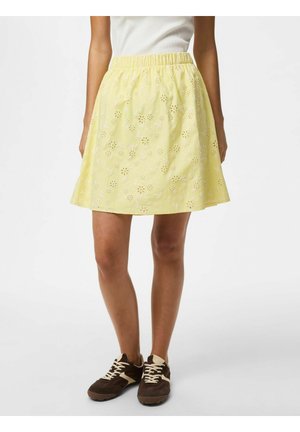 Kobieta w żółtej, kwiatowej spódnicy z oczkami i elastycznym pasem, białym topie oraz brązowych trampkach z białymi sznurowadłami, stojąca na białym tle.