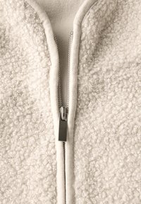 Veste en polaire de couleur blanc cassé, dotée d'un bord de tissu lisse et d'une fermeture éclair métallique à l'avant. Couche extérieure texturée avec une finition douce et en peluche.