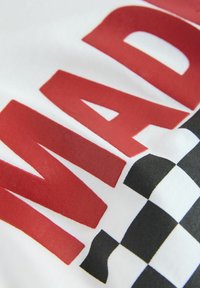 Gros plan sur les lettres rouges « MAD » et un motif à damiers noir et blanc sur un tissu blanc, probablement une partie d'un vêtement imprimé.