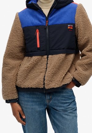 Personne portant une veste polaire beige avec un haut en color block bleu et noir et un détail de fermeture éclair corail, associée à un jean bleu et un col roulé beige.