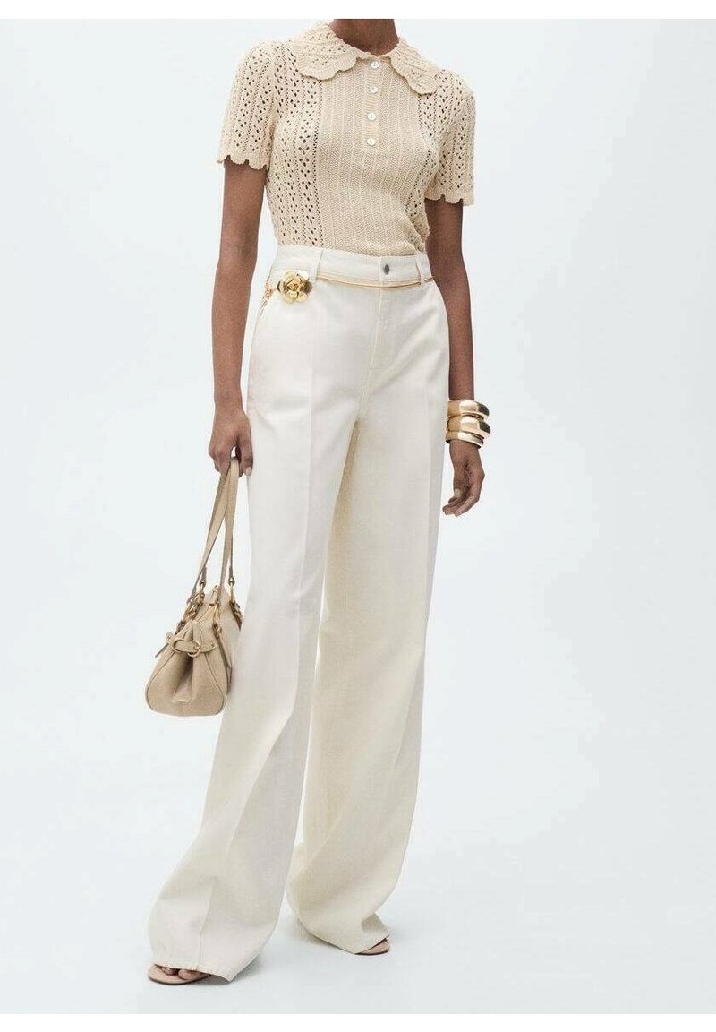 Beżowy sweter z dzianiny z falbaniastym kołnierzem, z przodu na guziki; białe szerokie spodnie z pasem w złotym kolorze, niosąc małą beżową torbę.