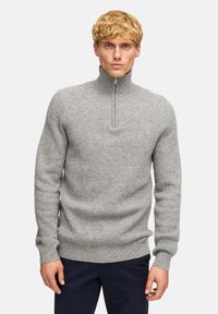 Maglione grigio lavorato a maglia con colletto alto e zip a un quarto. Tessuto a costine e maniche aderenti. Indossato sopra pantaloni blu navy.