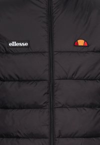 Svart puffer-väst med dragkedja, med horisontellt quiltade sektioner. Den har en vit "ellesse"-logotyp och ett litet flerfärgat emblem.