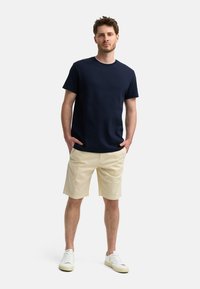T-shirt en coton bleu marine à col rond avec des manches courtes, associé à un short beige. Le mannequin porte des baskets blanches, les mains dans les poches.