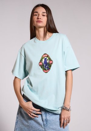 Camiseta de algodón azul claro con un colorido emblema bordado en el centro, combinada con una falda de mezclilla con un patrón de olas.