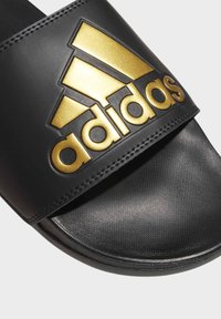 Černé skluzavkové sandály s lesklým černým páskem, na kterém je zlaté logo Adidas. Stélka je strukturovaná pro lepší přilnavost a podešev je mírně zakřivená.