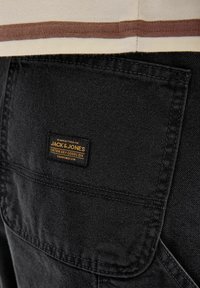 Crni traper s teksturiranom površinom, s oznakom "JACK & JONES" u narančastoj boji, koji je ušiven na stražnjem džepu.