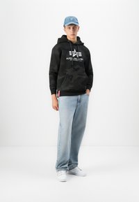 Joven vistiendo una sudadera con capucha de camuflaje negra de Alpha Industries, jeans de color azul claro, zapatillas blancas y una gorra de béisbol azul, de pie contra un fondo blanco.