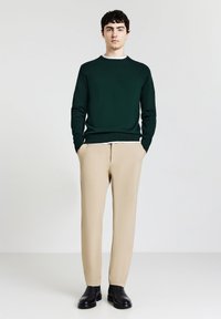 Grüner Strickpullover, beige Hosen und schwarze Lederschuhe. Einfaches Design, lockere Passform, glatte Textur und Kontrastelemente am Ausschnitt.