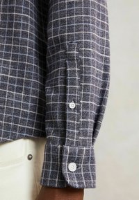 Cămașă de flanel în carouri bleumarin și alb, cu mânecă și manșetă cu nasture. Țesătură texturată, evidențiind un model de plasă proeminent și detalii netede ale nasturilor.