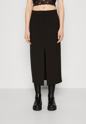 Femme portant une jupe midi noire taille haute avec une fente devant, un top court en dentelle noire et des bottes noires montantes et épaisses.