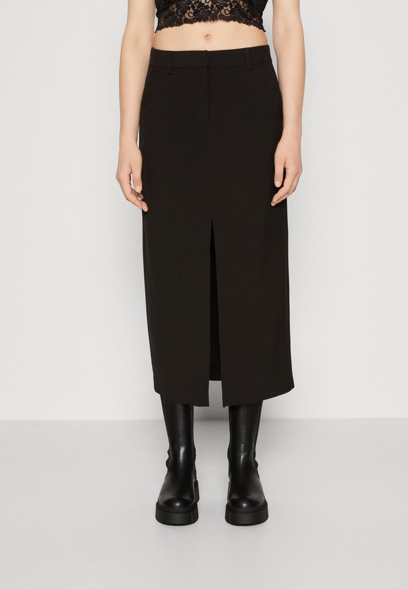 Femme portant une jupe midi noire taille haute avec une fente devant, un top court en dentelle noire et des bottes noires montantes et épaisses.